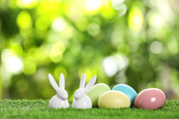 Ceramic Easter bunnies and dyed eggs on green grass against blurred background. Space for text