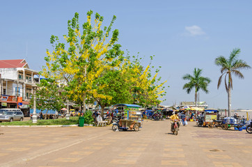 Kampong Chhnang City on the bank of the Mekong River with blossoming trees, mopeds, cars and a street vendor