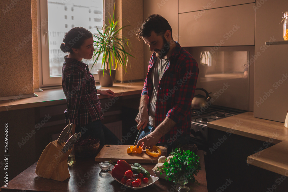 Wall mural beautiful young couple in kitchen at home while cooking healthy food. man cuts a pepper. woman looks