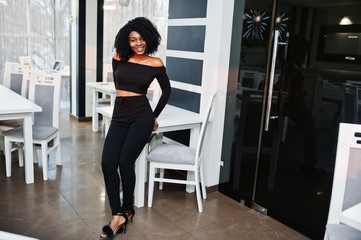 Fashionable young beautiful african american business woman with afro hairstyle wear in elegant black, stand at office near tables.
