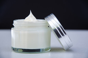 open glass jar of cream with a shiny lid on.white table. Black background.