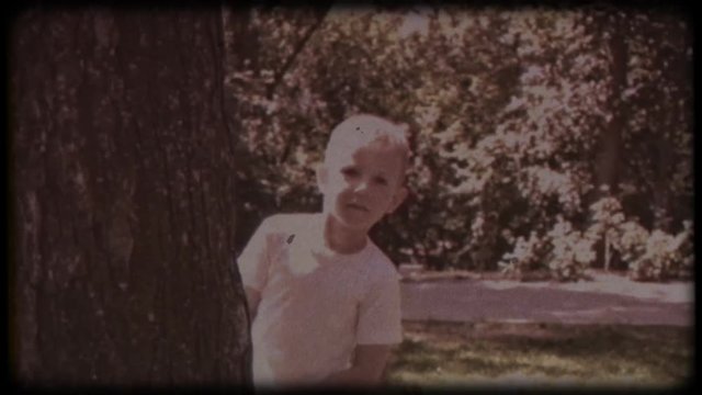 Family video archive. Retro camera 8&nbsp;mm. Old film. A blonde boy peeks out from behind a tree, playing hide and seek on a sunny summer day
