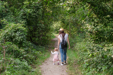 Beautiful brunette female in straw hat jeans, white t-shirt and a small backpack with his little cute child daughter playing together in a park