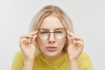 Portrait of a beautiful young woman with spectacles, looking curiously at you, squinting, trying to look closer. Girl with poor eyesight in yellow T-shirt posing on a white background in the studio - obrazy, fototapety, plakaty