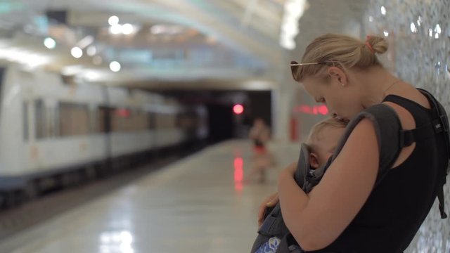 Mum with baby daughter waiting for train in subway. Woman kissing child sleeping in baby carrier