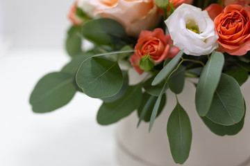 Delicate floral arrangement of roses (white, pink, orange) and green eucalyptus leaves in a round white cardboard box on a light background
