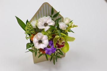 Wonderful floral arrangement of fresh flowers in a box in the form of an envelope on a light background. (Flowers: Falinopsis, eustoma, anemone, leaves. Colors: white, green, yellow).
