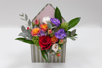 Original floral arrangement (gift) of fresh flowers in a box in the form of an envelope on a light background. (Flowers: roses, eustoma, leaves. Primary colors: red, purple, orange, white, green).