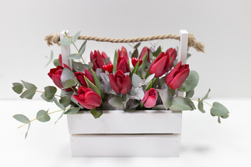A stylish arrangement of fresh flowers (red tulips and green eucalyptus leaves) in a wooden box of white color with a rope handle