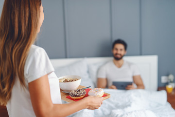 Obraz na płótnie Canvas Attractive brunette bringing breakfast to her loving husband. Man sitting in bed and using tablet. Selective focus on woman.
