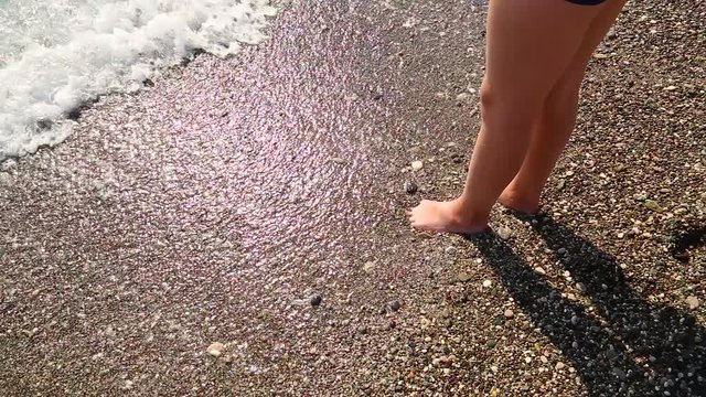 View from above cute wet legs of young kid standing at sunny summer beach and enjoying clear warm sea water. Boy walks into water and shadow of body appears on sea surface. full hd video footage.