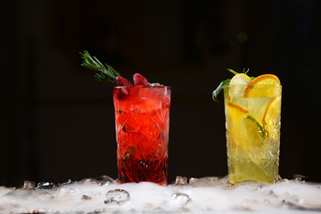 Two tall crystal glasses with lemonade made from orange, lime, strawberries, blueberries and raspberries on a black background and pieces of ice on the table. Nitrogen vapor in the frame.