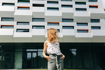 Beautiful hipster girl is standing near the modern architecture building. Stylish business woman walking near the fashionable lines, glass and windows. Green grass lawn. Office worker break.