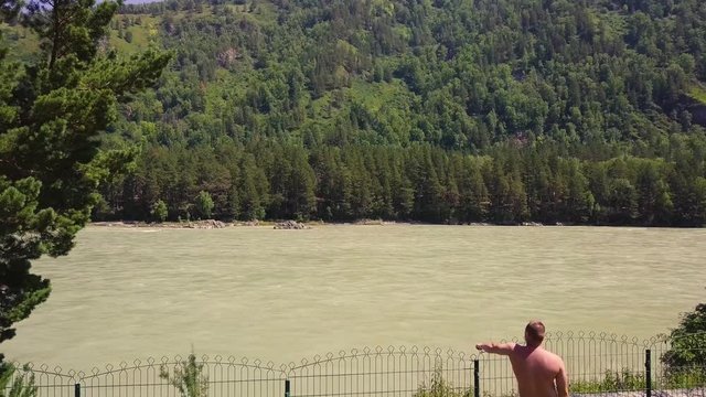 Aerial: Flying over a mountain river with a rapid current. A man dressed in panties looks at the river, showing his hand. Also visible: river rapids, pine forest.
