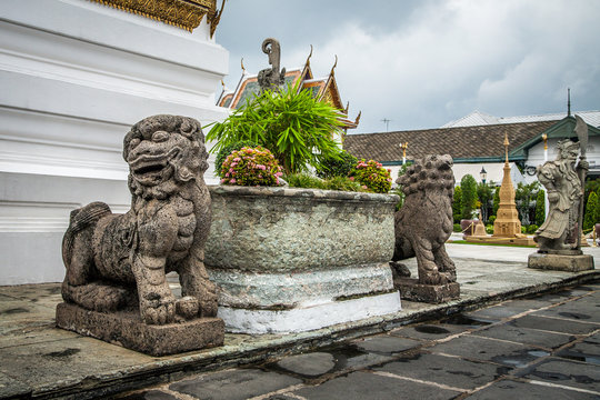 Chinese dog or lion sculpture near flowerpot with flowers, pagodas with guardian on a background, in Grand Palace Bangkok, Thailand