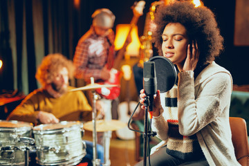 Mixed race woman singing. In background band playing instruments. Home studio interior.