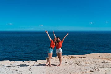 Two young back women friends with raised hands, looking at sea. Freedom concept, holiday, beach