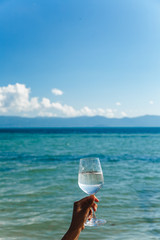 Aquamarine sea background with blue sky and white clouds skyline wine glass with brilliant liquid natural light tanned female hand and shadows and caustic landscape