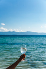 Aquamarine sea background with blue sky and white clouds skyline wine glass with brilliant liquid natural light tanned female hand and shadows and caustic landscape