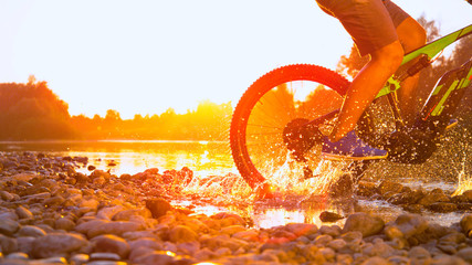 SLOW MOTION CLOSEUP Unrecognizable man MTB biking in shallow river at sunset - obrazy, fototapety, plakaty