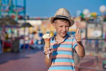 Smiling boy with the hat on his head is standing in the amusement park with the ice-cream in his hands.