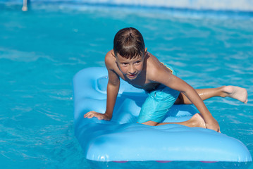 Cute European boy is enjoying his summer holidays. He is having fun with the blue inflatable air mattress in the hotel’s swimming pool.