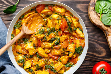 Top view on Bombay chicken curry with spinach, tomato and onion in a white frying pan on a kitchen wooden table. Indian traditional cuisine.
