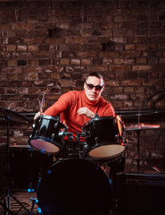 Fototapeta na wymiar Young stylish musician in sunglasses sits behind the drum set against a brick wall, thoughtfully looking at the camera. Perform in a night club