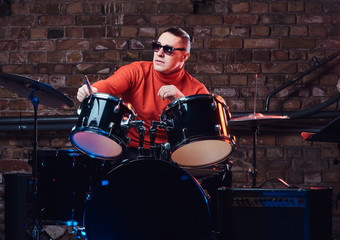 Fototapeta na wymiar Young stylish musician in sunglasses sits behind the drum set against a brick wall, perform in night club