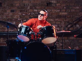 Fototapeta na wymiar Young stylish musician in sunglasses sits behind the drum set against a brick wall, perform in night club