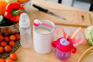 Close-up of dried milk, bottles for preparing baby milk formula on wooden table. Soluble milk powder in dosing spoon. Healthy fresh foods are on the table in the kitchen