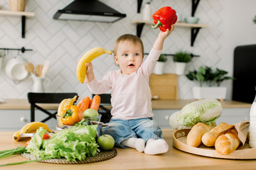 Fototapeta premium Cheerful baby child holds banana and red pepper while sitting on a table on the background of a modern light kitchen