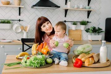 mother and little kid preparing healthy food and having fun. Baby girl sitting on the wooden table with fresh vegetables and fruits and holds apples