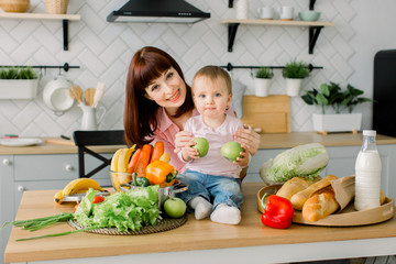 mother and little kid preparing healthy food and having fun. Baby girl sitting on the wooden table with fresh vegetables and fruits and holds apples