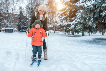 Fototapeta na wymiar A young woman, mom supports with care, skiing in winter in the forest park, a little son a boy of 3-6 years. The first sports lessons on the street. Caring for parent-child. Free space for text.