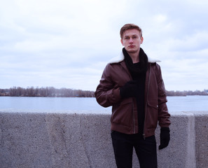 A young blond man in a leather jacket on the promenade near the river in the city.