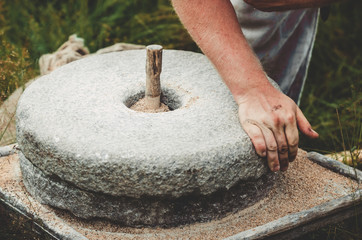 The ancient quern stone hand mill with grain. The man grinds the grain into flour with the help of a millstone. Men's hands on a millstone. Old grinding stones turned by hands © marmoset