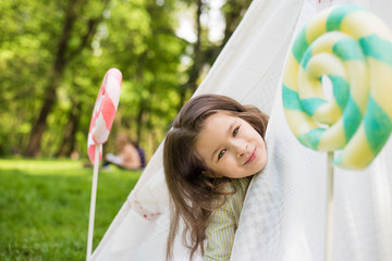 Little girl lying and playing in a tent, children's house wigwam in park. Happy mothers day. Summer, childhood concept. Big lollipop © MartaKlos