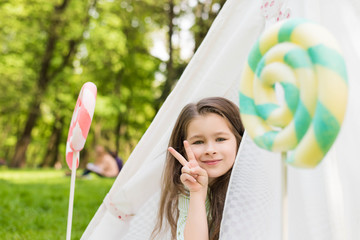 Little girl lying and playing in a tent, children's house wigwam in park. Happy mothers day. Summer, childhood concept. Big lollipop