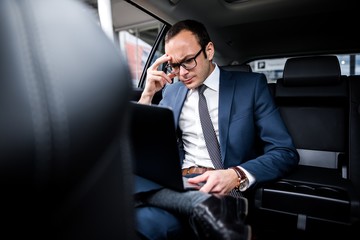 Brooding businessman with glasses sitting in the back seat of a car working on a laptop solving problems