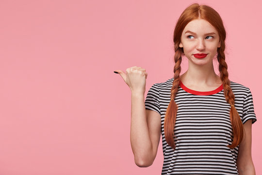 Red-haired girl with two braids pointing with thumb to the left side on blank copy space and looks there mysterious intriguing, smiling playfully, conceived something, on pink wall