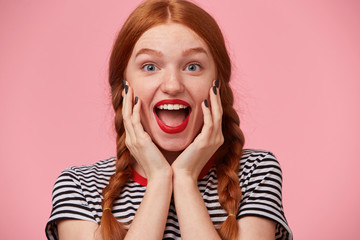 Overflowing with positive emotions joyful red-haired girl with two braids keeps hands near her face and openes mouth widely in excitement, with red lips, white healthy teeth, isolated on pink wall