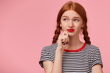 Unsatisfied frowning red-haired girl with two braids keeps fist near the chin and looking to the upper left corner without desire,enthusiasm,with discontent, isn't thrilled with new idea, on pink wall