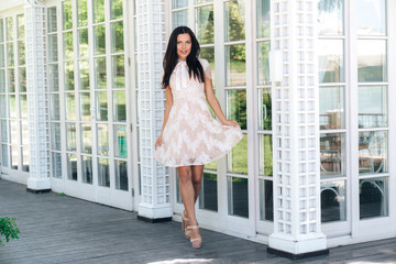 Cute brunette with beautiful long legs posing in beige colour dress outside near a wooden and glass wall