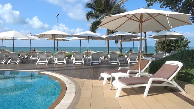 resort pool area with sun chairs under white umbrellas in foreground, turquois ocean, blue sunny sky with cumulus clouds, locked shot afternoon daylight scenery, no people
