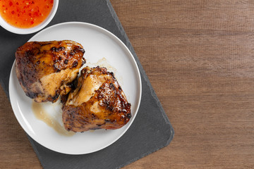 top view of grilled chicken thigh in a ceramic dish served with sweet chilii sauce on wooden table. homemade style food concept.