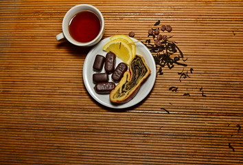 A cup of tea candy in a plate with lemon a slice of bun next to sprinkled tea on a light cane background copy space