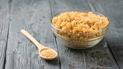 Wooden spoon with quinoa and a glass bowl with the cooked quinoa on a black wooden table.