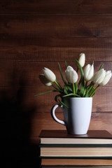 Bunch of white tulips in mug on stack of books against wooden background. Spring concept. Springtime, love reading, library, education