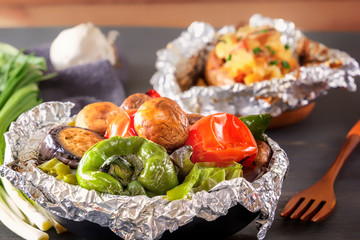 Baked vegetables in foil - tomatoes, eggplants, peppers and baked potatoes with bacon, onions on a gray wooden table. Close-up